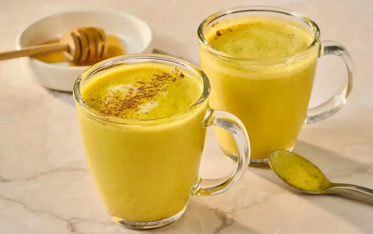 Zwei Tassen goldgelbe Milch mit Kurkuma und Honig, garniert mit Zimt. Ein Honigl&ouml;ffel ruht in einer Schale mit Honig.