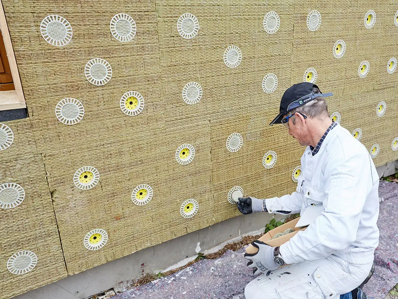 Pracownik montuje kołki do wełny mineralnej na elewacji. Widać żółte elementy mocujące.