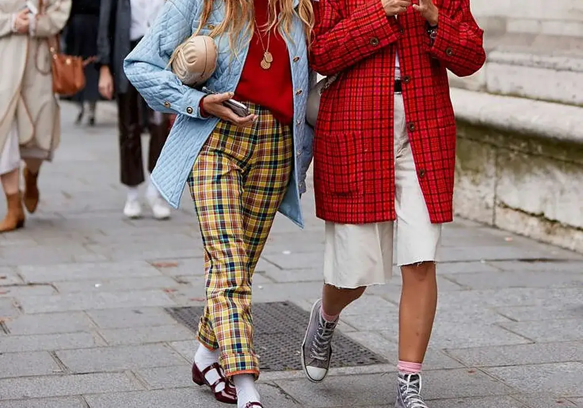 Dos mujeres pasean con un estilo cayetano audaz: una con pantalón de cuadros amarillos y chaqueta azul, la otra con falda blanca y chaqueta roja a cuadros.