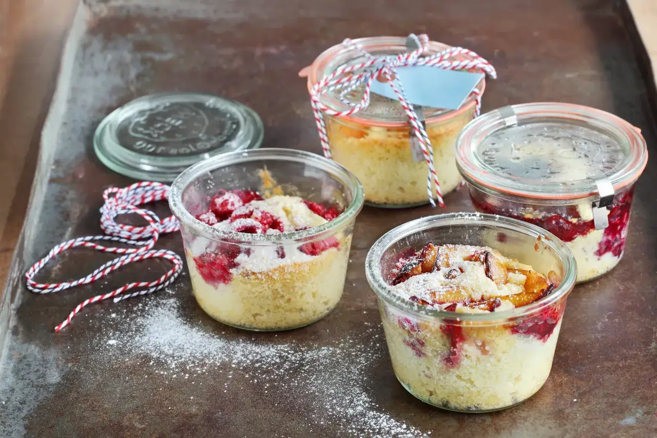 Vier leckere Kuchen im Glas, mit Himbeeren und Pflaumen, bestäubt mit Puderzucker.