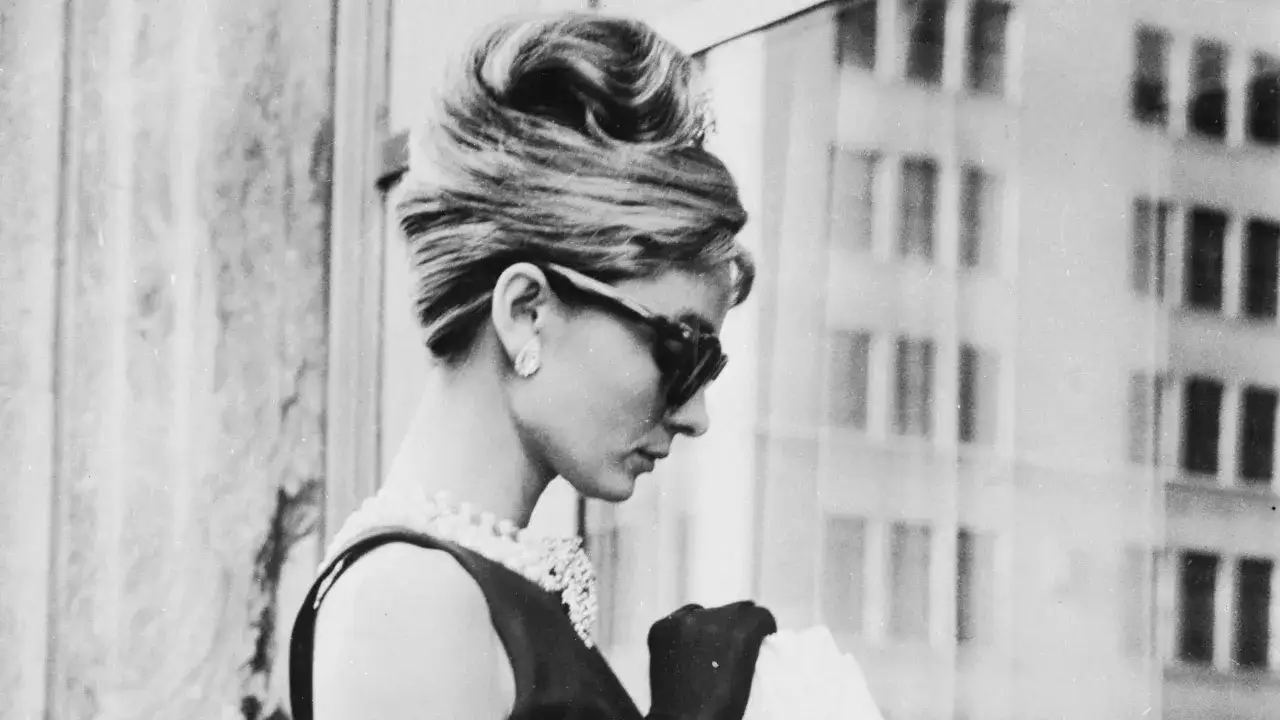 Audrey Hepburn, con su icónico peinado y gafas de sol, luce elegante con perlas y guantes negros.