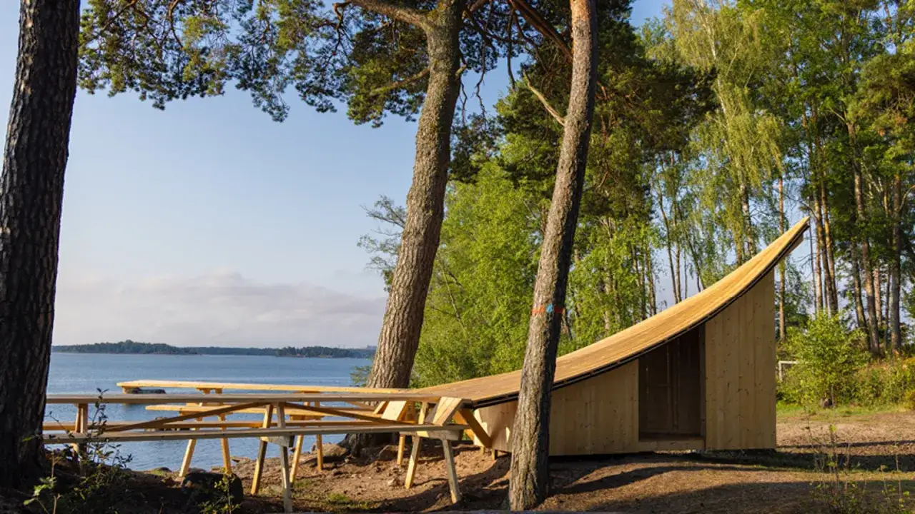 En vacker plats f&ouml;r yoga utomhus vid en sj&ouml;. En tr&auml;byggnad med sv&auml;ngd taklinje och picknickbord i f&ouml;rgrunden.