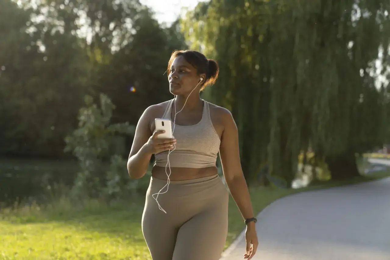 Femme souriante marchant dans un parc avec son téléphone et des écouteurs, profitant du soleil. Une belle façon de perdre du poids en marchant.