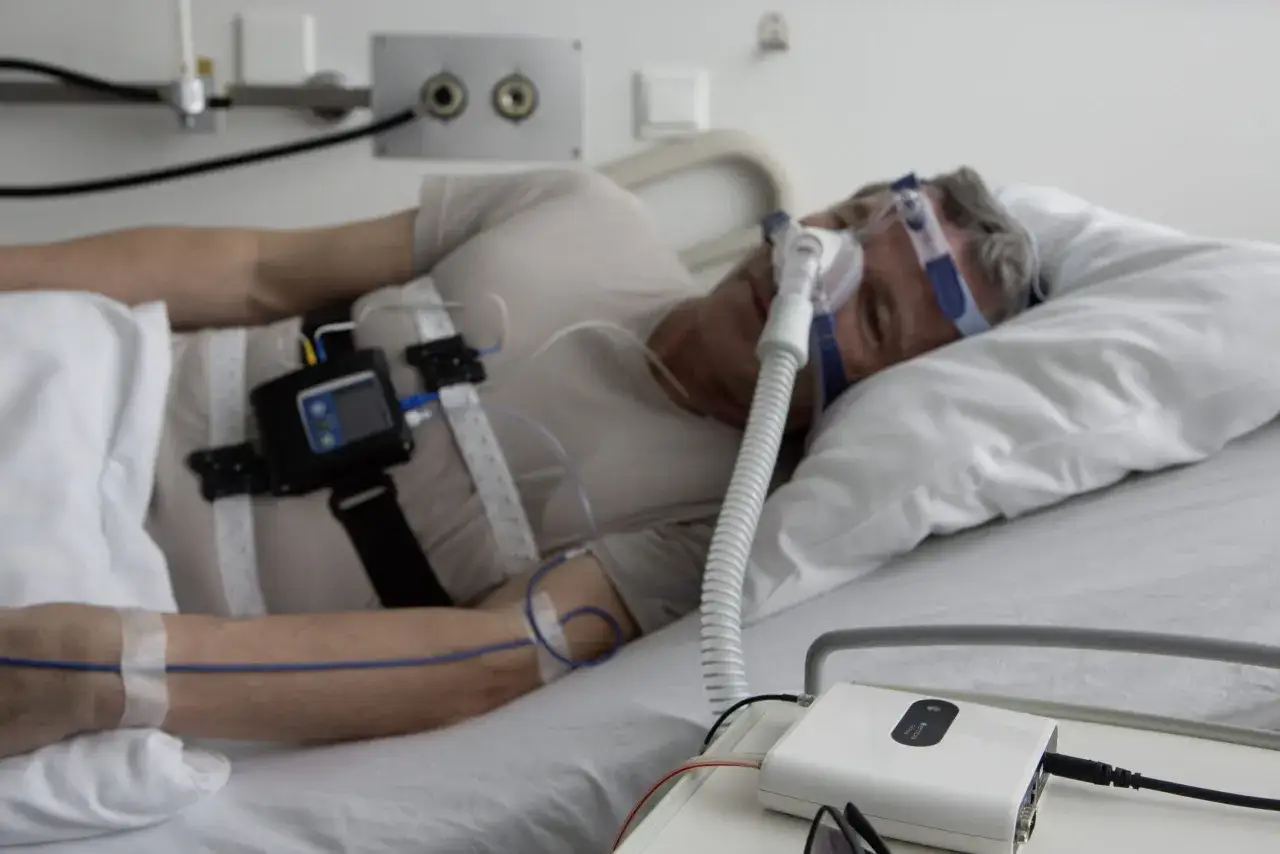 Mężczyzna w masce CPAP śpi w ł&oacute;żku szpitalnym, podłączony do aparatury monitorującej. Czy to płakanie przez sen?