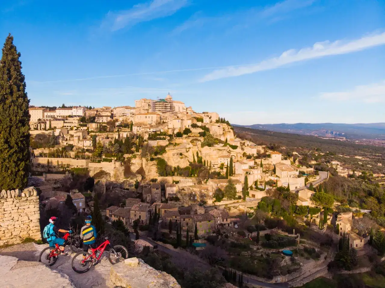 Deux cyclistes contemplent la ville de murs perch&eacute;e sur une colline, sous un ciel bleu.