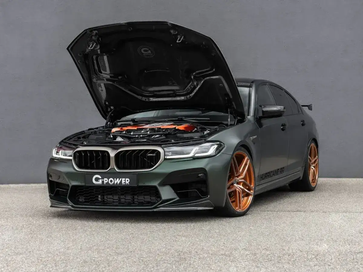 Mocarny BMW M5 G Power z otwartą maską, odsłaniającą pomarańczowy silnik. Masywne, miedziane felgi podkreślają jego sportowy charakter.