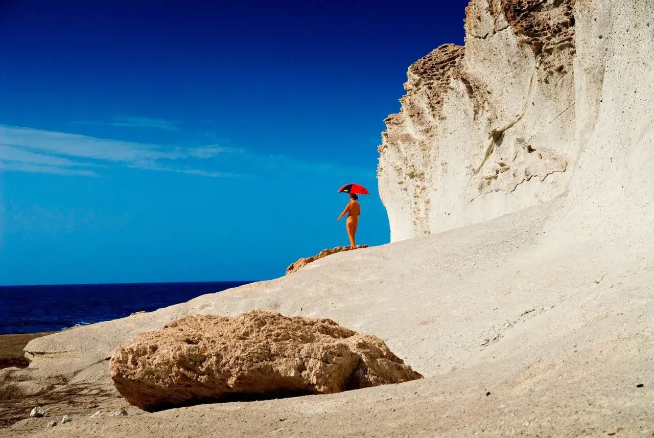 Figura solitaria con sombrilla roja en las playas nudistas de Cabo de Gata, bajo un cielo azul intenso y acantilados blancos.