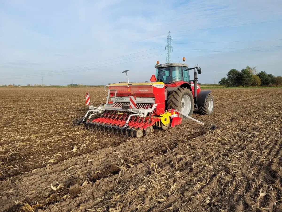 Czerwony traktor z siewnikiem wykonuje uprawę bezorkową na polu. Maszyna rozsiewa nasiona, przygotowując glebę pod nowy zasiew.