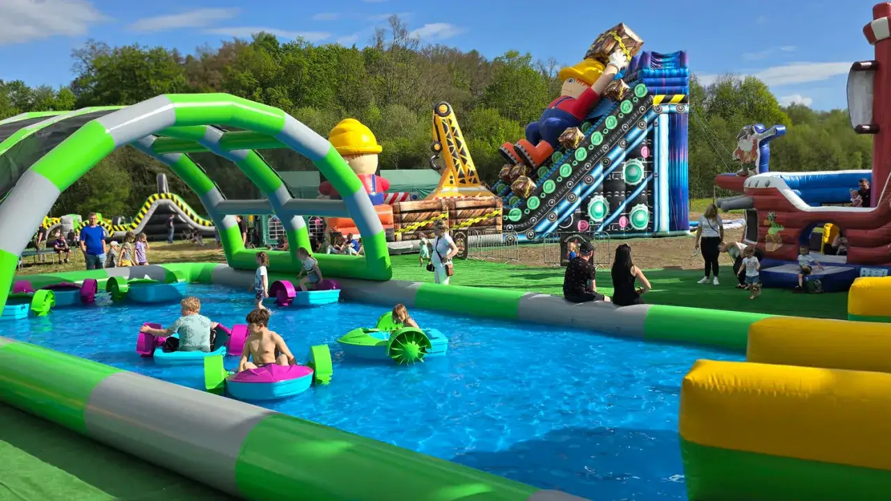 Wesoły park dmuchańców z basenem, zjeżdżalniami i dmuchanymi łódkami. Dzieci bawią się w wodzie, a na tle widać kolorowe dmuchane konstrukcje.