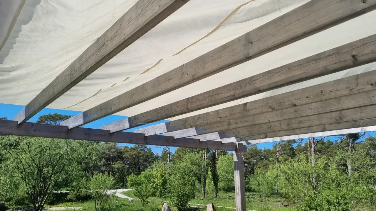 Tr&auml;bj&auml;lkar st&ouml;der ett vitt pergolatak av tyg, som ger skugga &ouml;ver en gr&ouml;nskande tr&auml;dg&aring;rd med bl&aring; himmel.