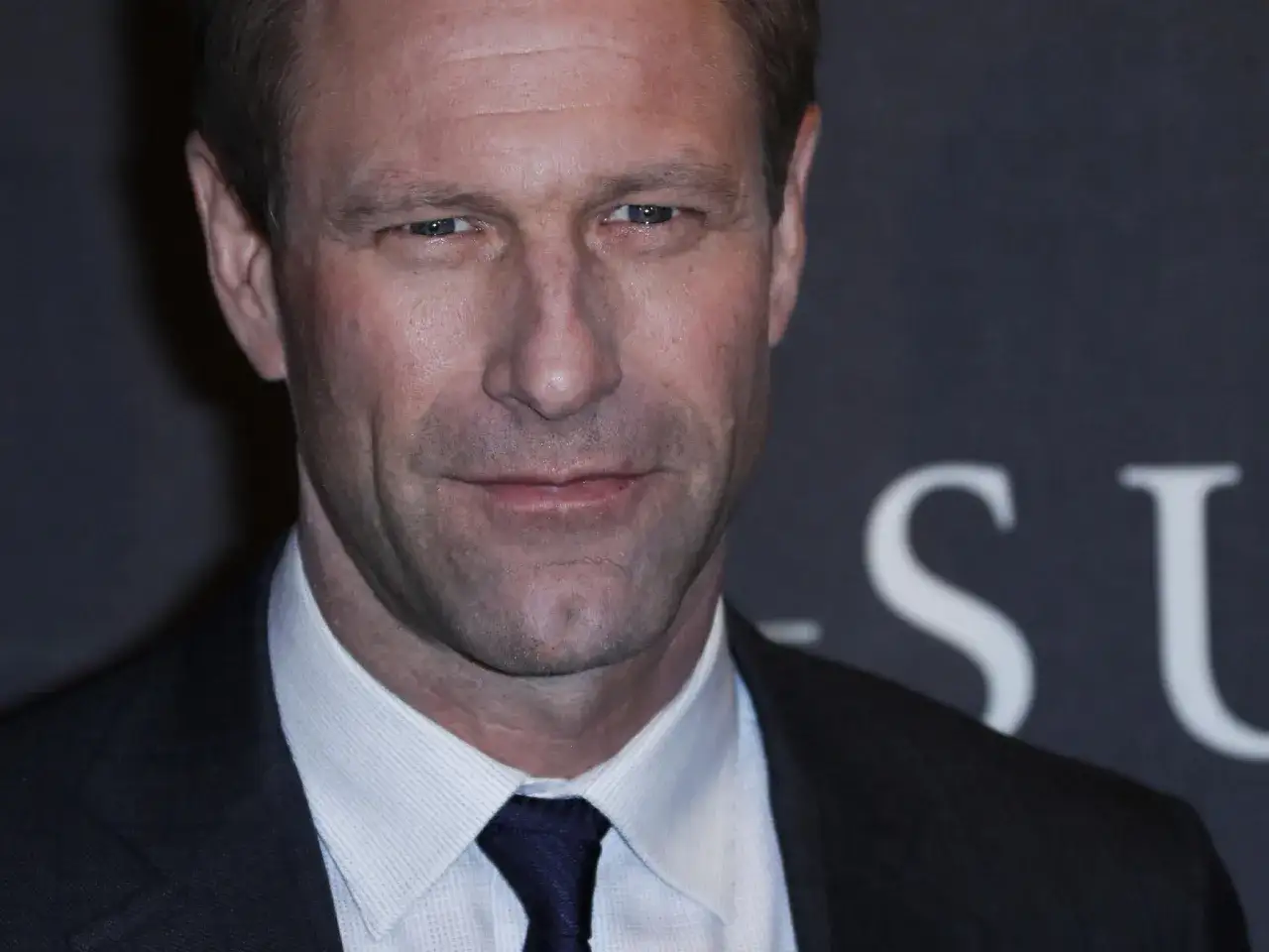 Primo piano di Aaron Eckhart, attore noto per i suoi ruoli in film intensi. Indossa una camicia bianca e una cravatta blu scuro.