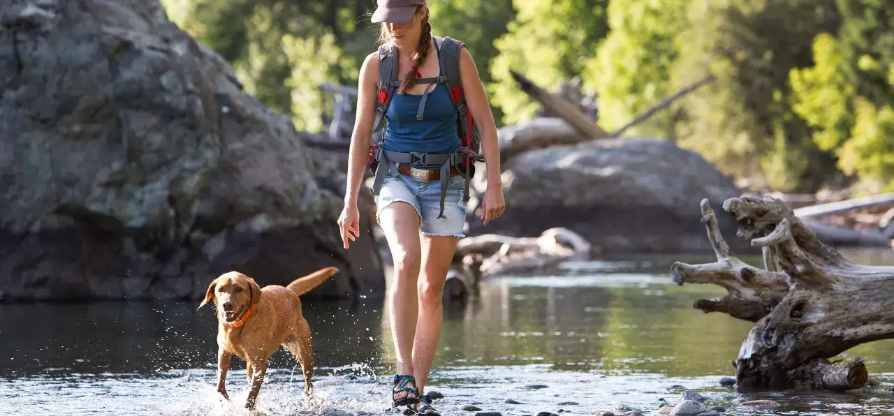 Une femme avec un sac à dos et son chien traversent une rivière. Une activité insolite avec son chien pour se rafraîchir.
