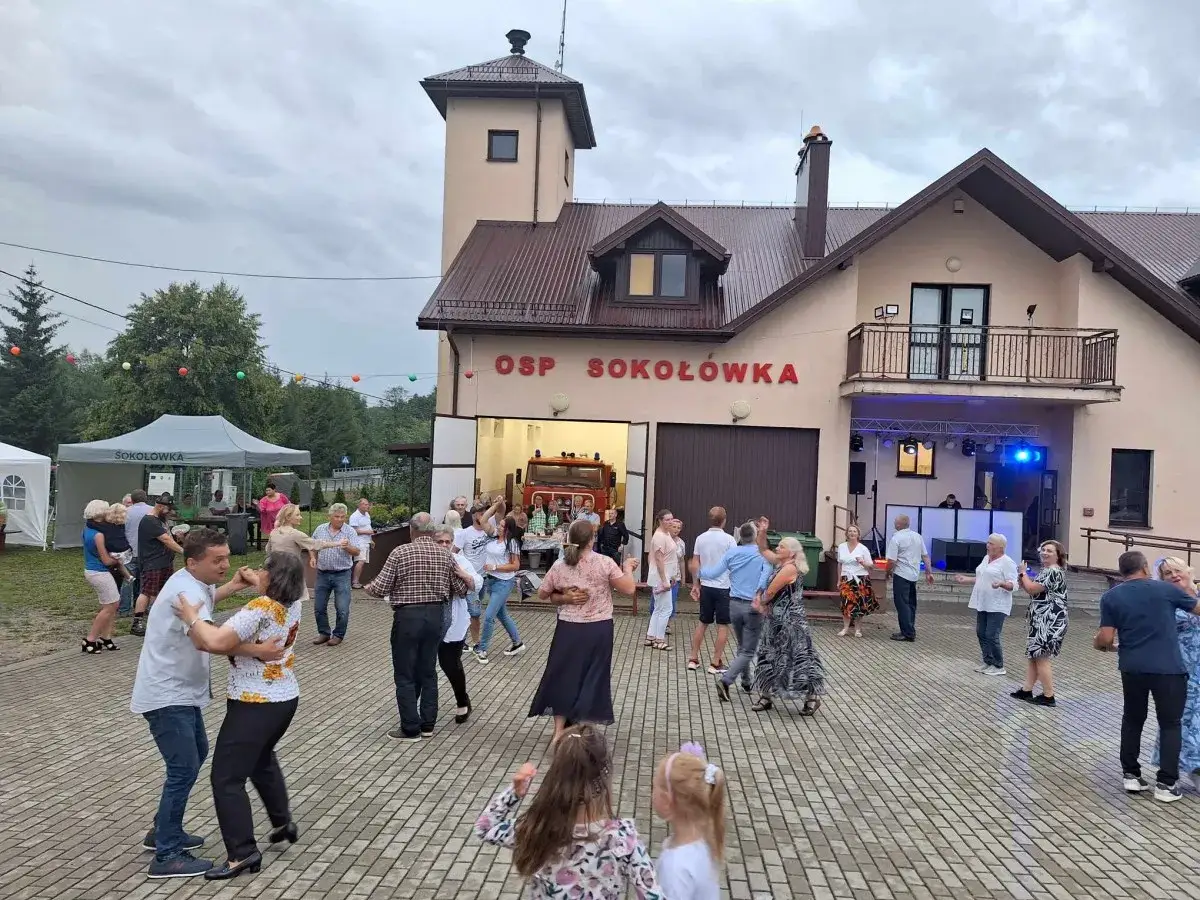 Ludzie tańczą przed remizą OSP Sokoł&oacute;wka. W tle widać w&oacute;z strażacki i scenę z DJ-em. Wszyscy świetnie się bawią, to prawdziwa zabawa w remizie!