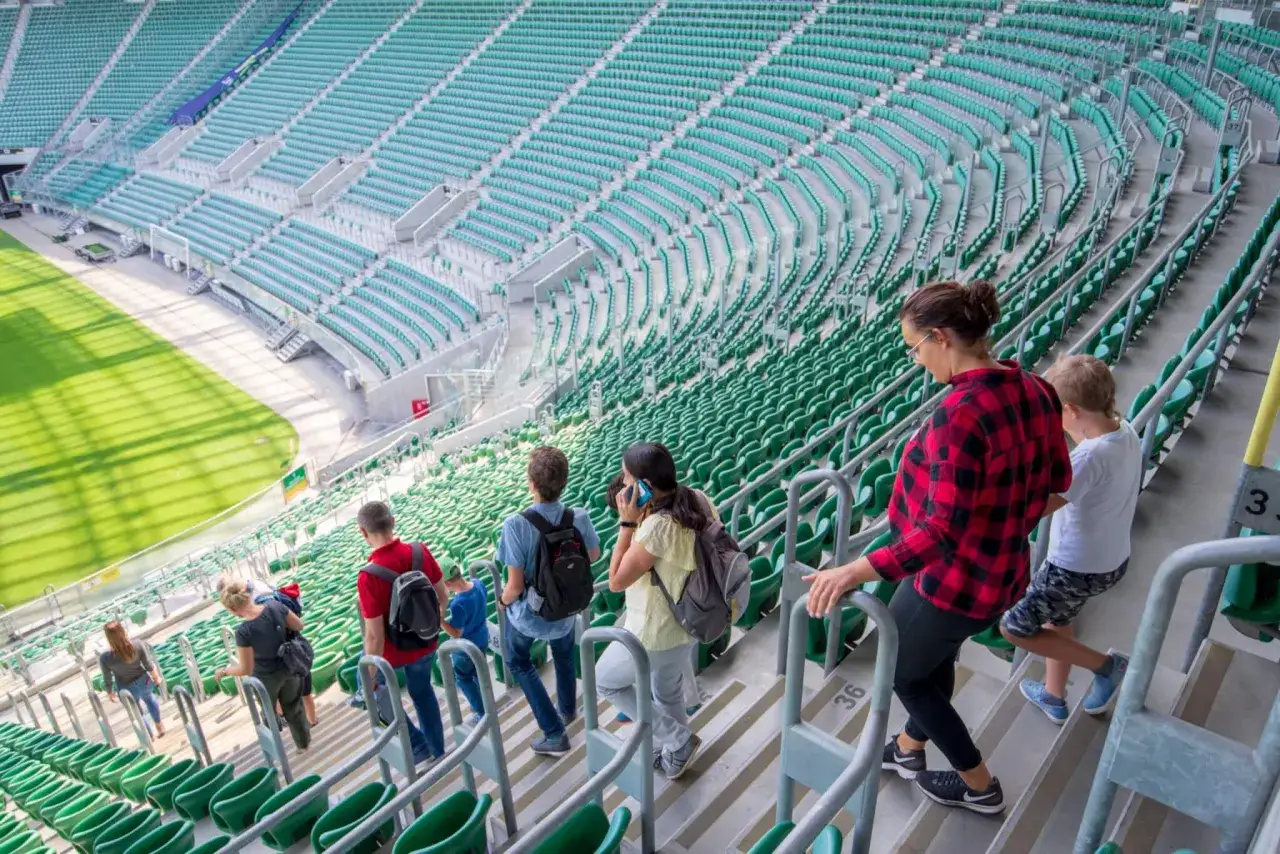 Grupa osób wchodzi na zielone trybuny stadionu. Tarczyński Arena pojemność robi wrażenie, widzimy rzędy pustych siedzeń.