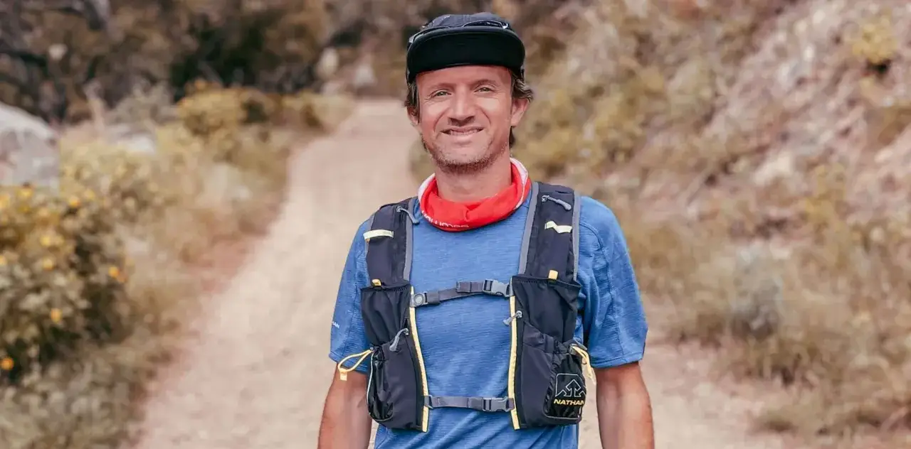 Un homme souriant, pr&ecirc;t pour une trail longue distance, porte un sac de trail et un bandana rouge. Le chemin s'&eacute;tend derri&egrave;re lui.
