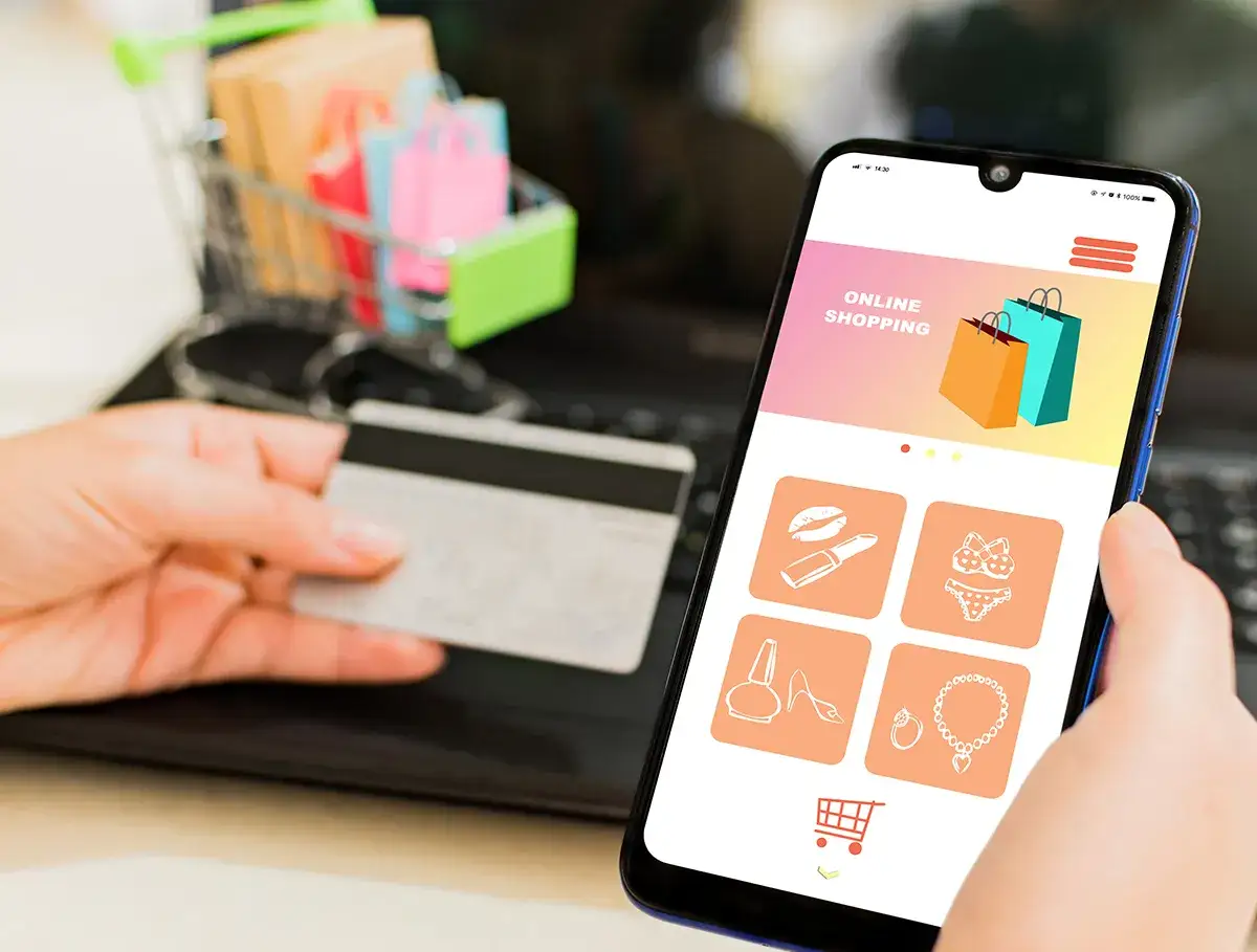 E-commerce deutsch: H&auml;nde halten Kreditkarte und Smartphone mit Shopping-App. Mini-Einkaufswagen mit T&uuml;ten im Hintergrund.