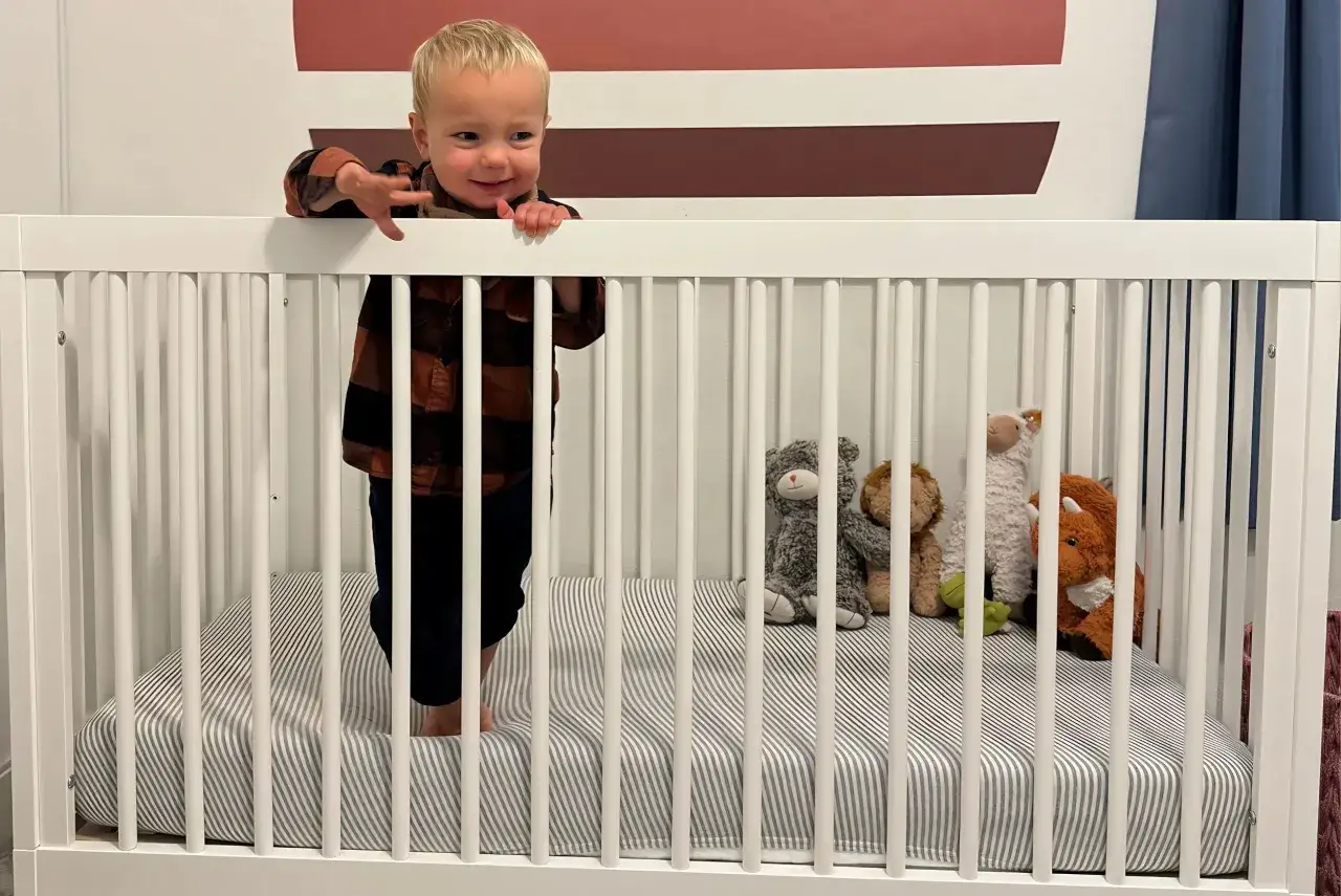 A happy toddler stands in a mini crib, surrounded by stuffed animals. This crib is designed for safety, with a clear mini crib weight limit.