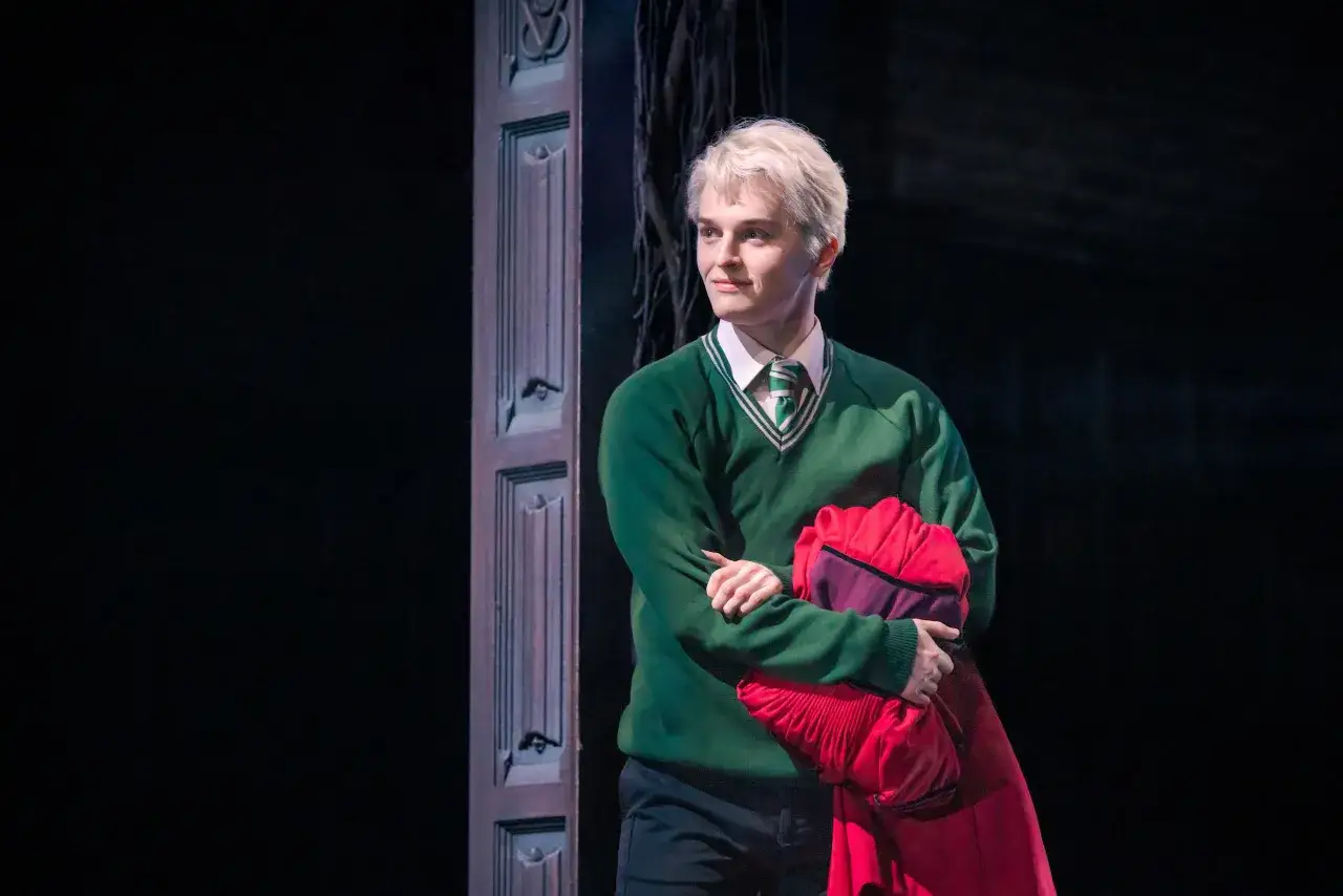 Scorpius Malfoy w zielonym swetrze i krawacie, trzymający czerwoną pelerynę, stoi obok ozdobnych drzwi.