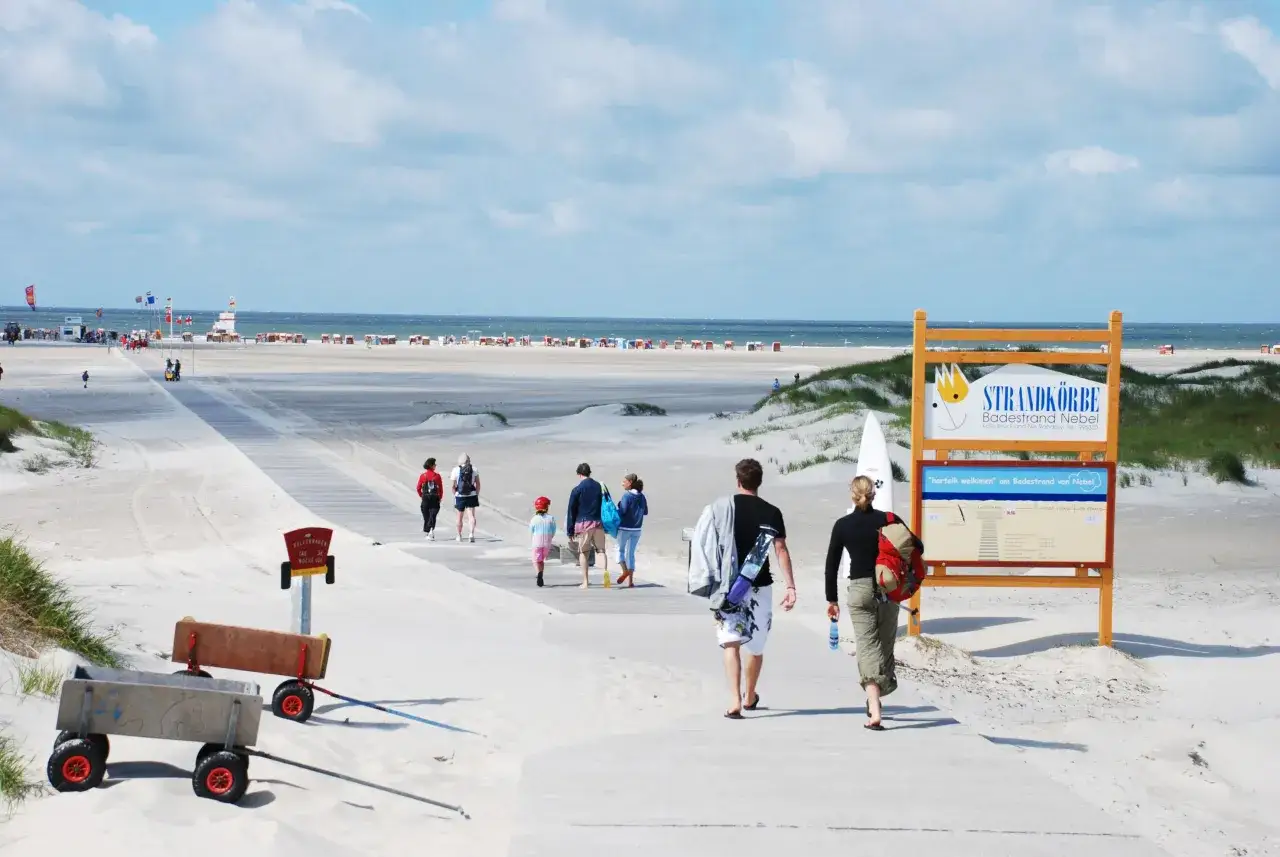 Menschen genießen das Leben und Arbeiten auf Amrum. Sie spazieren über den Holzsteg zum Strand mit Strandkörben und dem Meer im Hintergrund.