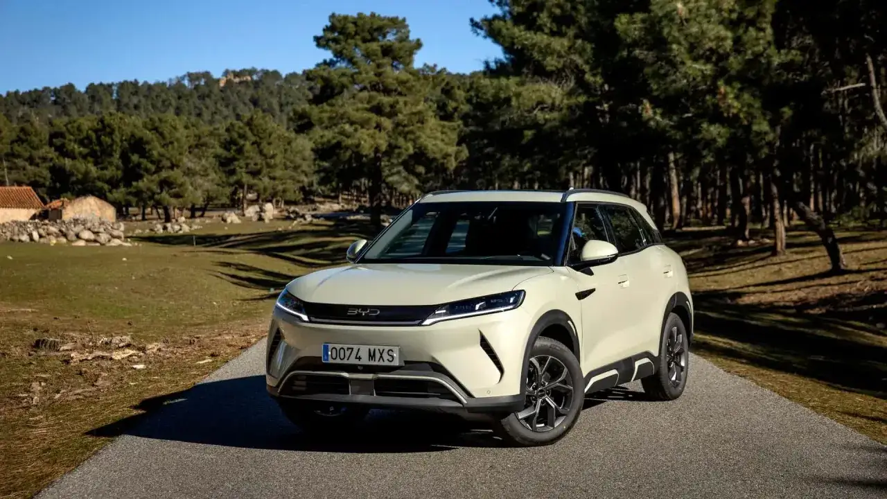 BYD Atto 3, un SUV elettrico color crema, parcheggiato su una strada di campagna con alberi e un edificio in pietra sullo sfondo. Scopri il byd atto 2 prezzo.