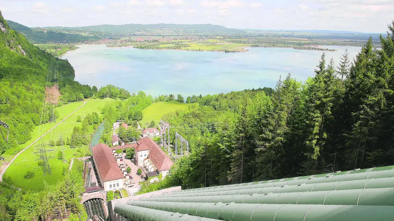Große Rohre führen zu einem Kraftwerk am See. Die Nutzung von Wasserkraft ist hier sichtbar, umgeben von grünen Hügeln und Bäumen.