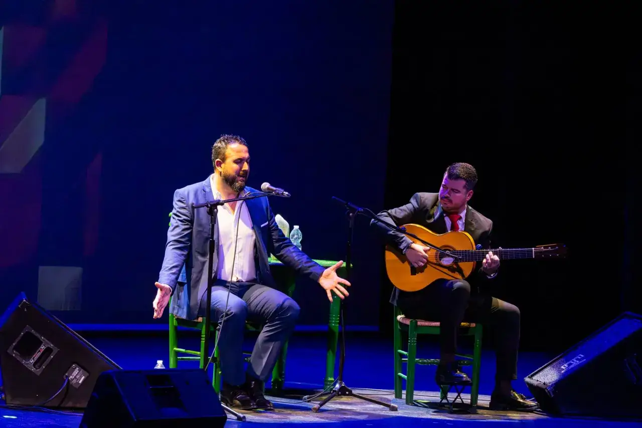 Dos cantaores flamencos famosos, uno cantando y otro tocando la guitarra, en un escenario con luces azules.