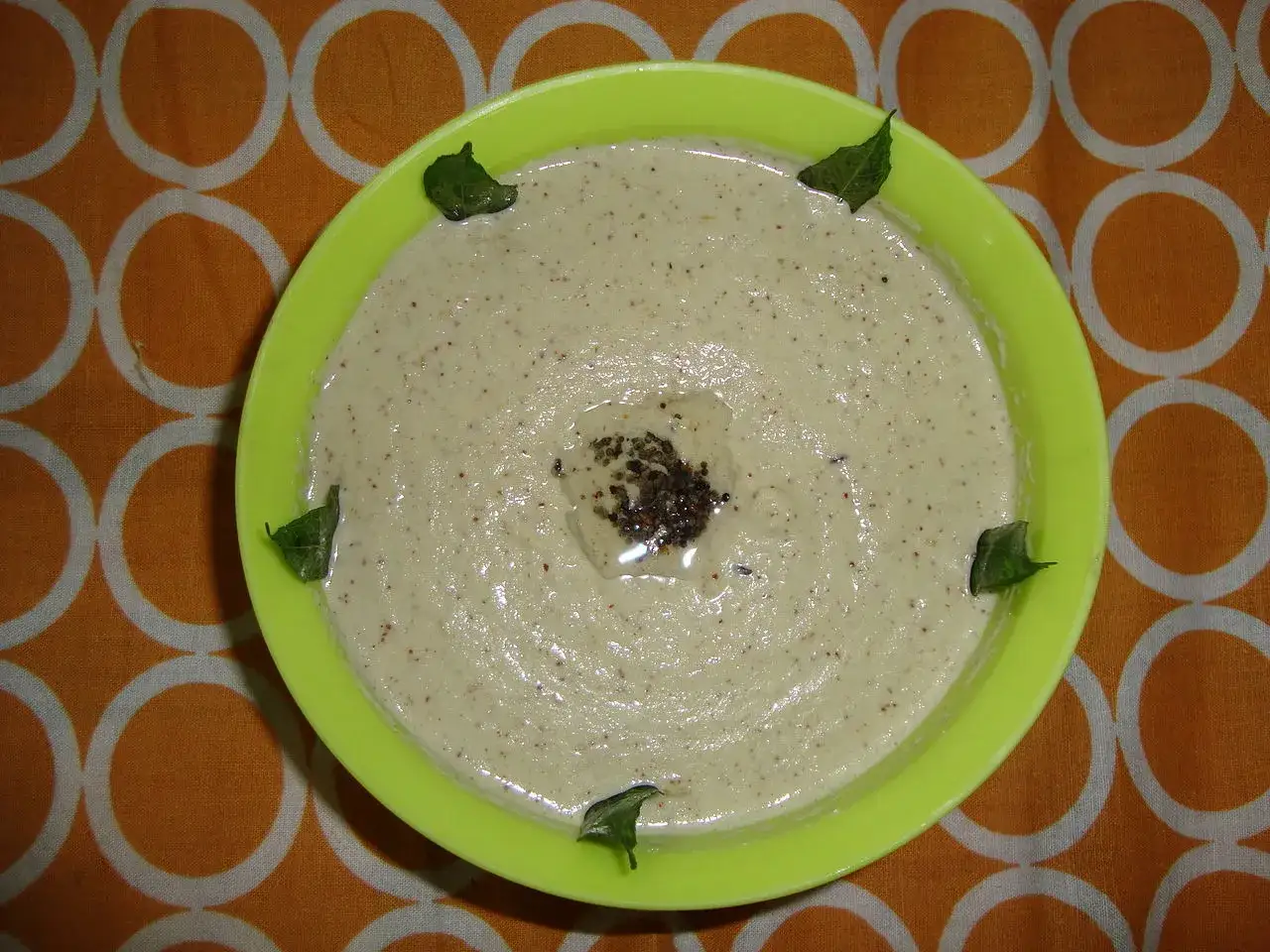 Kremowy sos z mleka kokosowego, udekorowany listkami curry i przyprawami, w jasnozielonej miseczce na pomarańczowym tle.