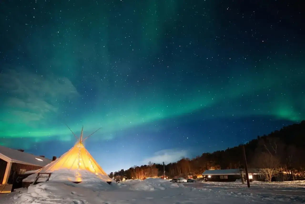 Nocne niebo nad **Nordkapp camping** rozświetla zorza polarna. W śnieżnej scenerii widać oświetlony namiot i domki.