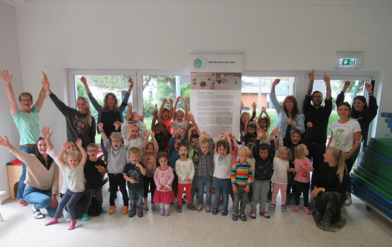 Fr&ouml;hliche Kinder und Erzieher*innen mit erhobenen Armen zeigen Beispiele f&uuml;r Demokratie im Kindergarten.