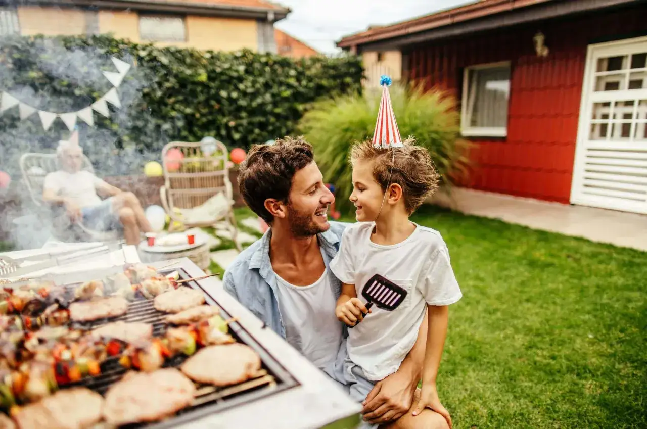 Vater und Sohn genie&szlig;en das Grillen mit Kindern im Garten. Rauch steigt vom Grill auf, auf dem Spie&szlig;e und Burger brutzeln.