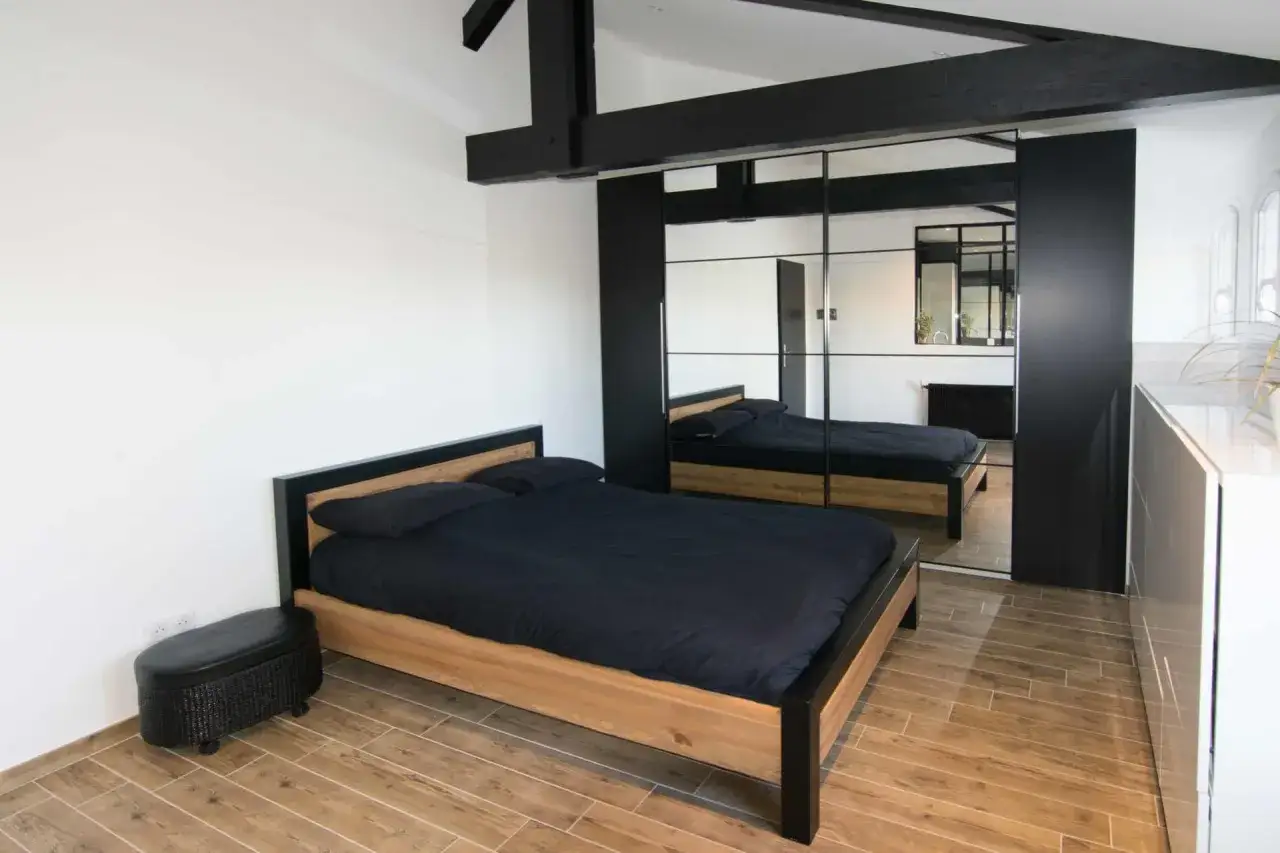 Une chambre moderne avec un lit en bois et des draps noirs, des poutres apparentes et une grande armoire miroir. Idéal pour refaire sa chambre avec style.