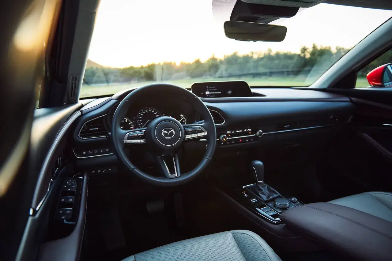 Interi&ouml;rbild av en Mazda CX-30 hybrid. Ratt, instrumentpanel och mittkonsol syns. Solnedg&aring;ng i bakgrunden.