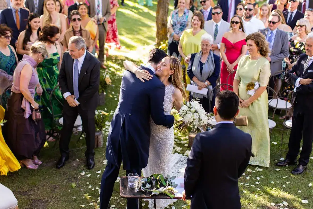 Una pareja se abraza en una boda al aire libre, rodeada de invitados. El novio, con chaqué, abraza a la novia, vestida de encaje. Las **canciones de boda modernas** seguro que amenizan este emotivo momento.