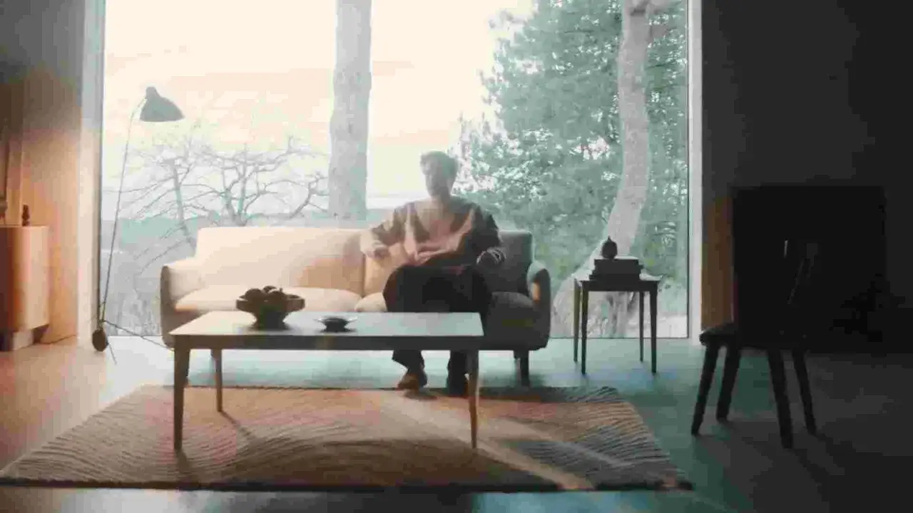 En person sitter i en soffa i ett ljust rum med stora fönster. Minimalistisk dansk inredning inspiration med en soffa, soffbord och matta.