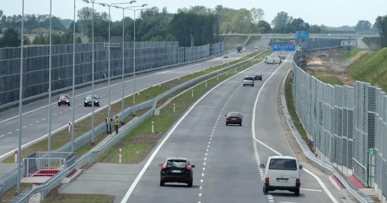 Samochody jadą lewym pasem autostrady, mijając ekrany akustyczne i drzewa.