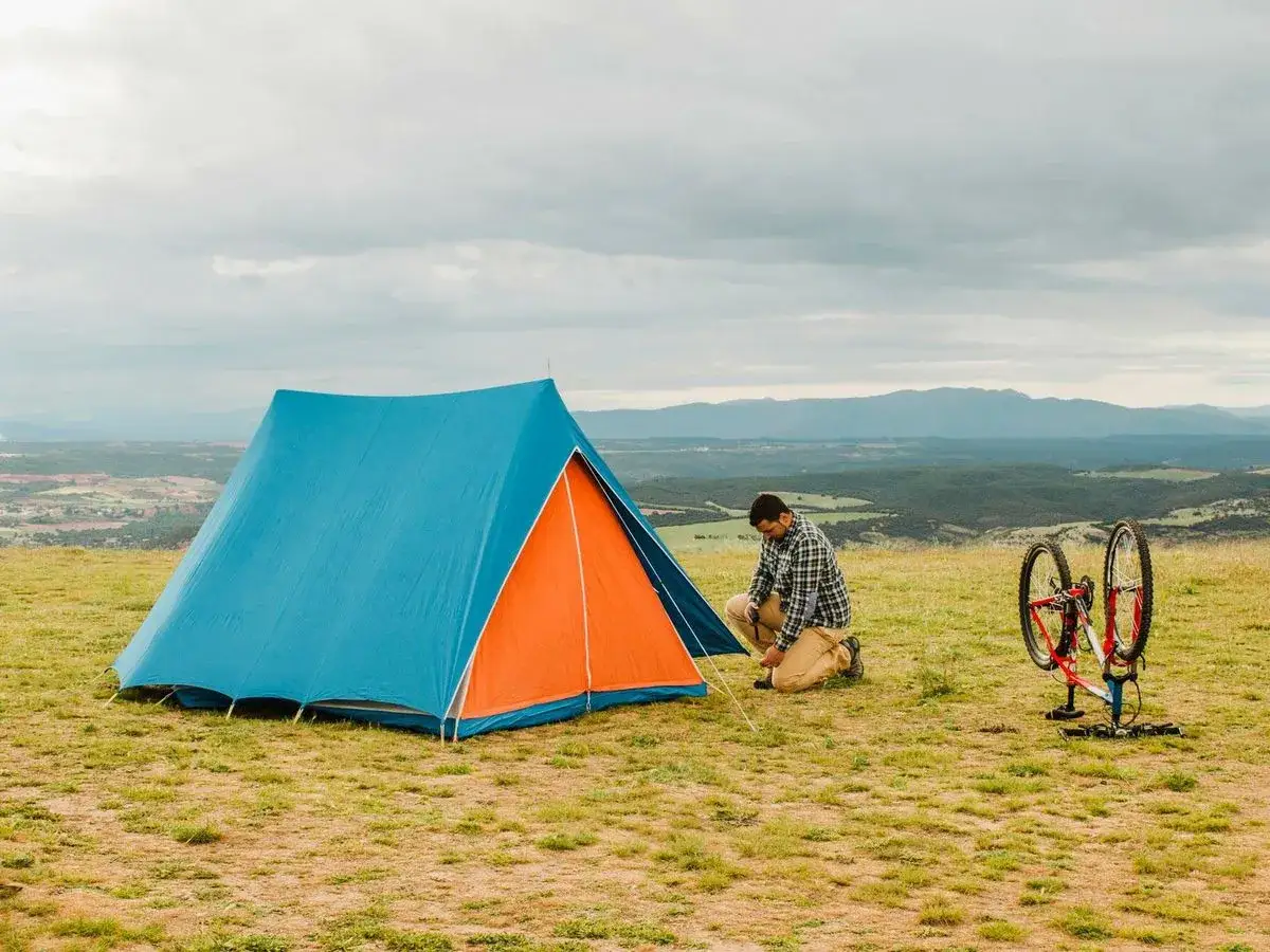 Hombre montando una tienda de campa&ntilde;a azul y naranja en la cima de una colina, listo para como acampar. Bicicleta a un lado.