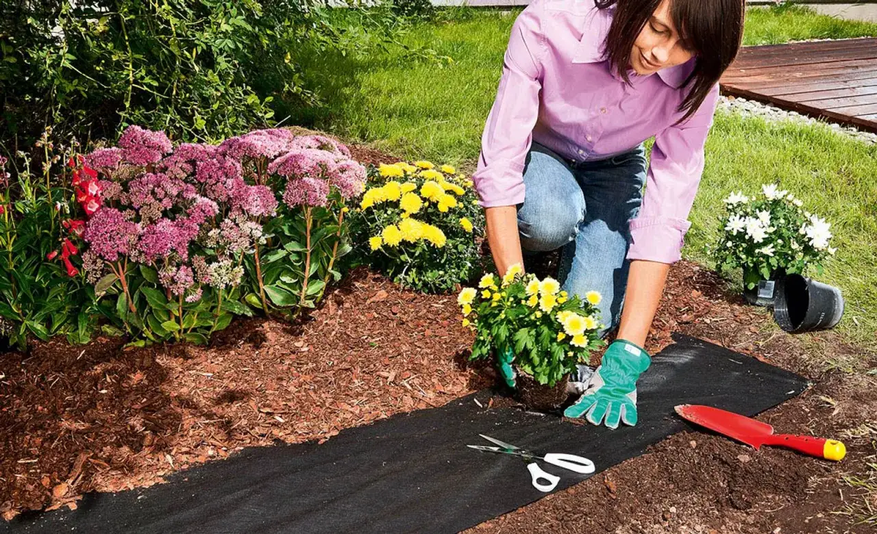Frau verlegt Unkrautvlies im Garten, um neue Blumen zu pflanzen. Schere und Spaten liegen bereit.