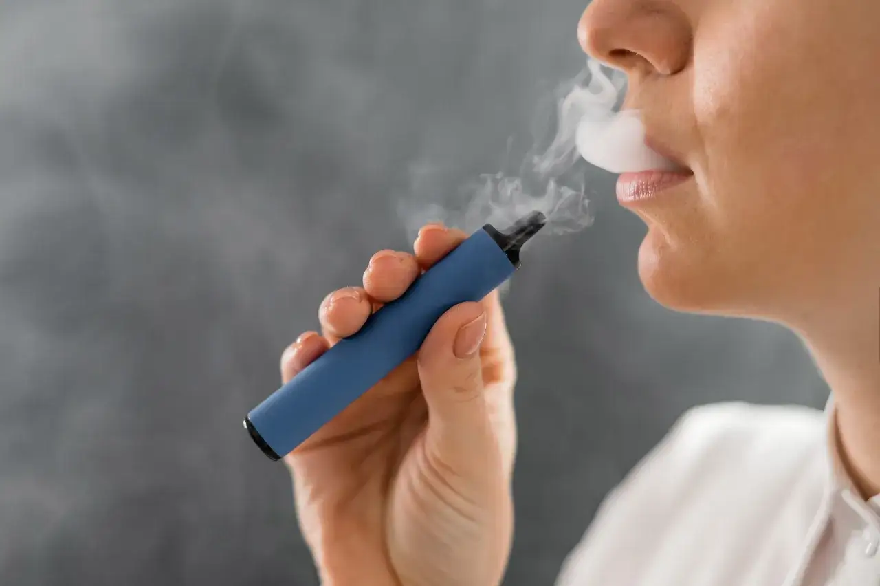 Persona vapeando exhala humo de un dispositivo azul. La imagen plantea la pregunta sobre cuanto vapear al dia.