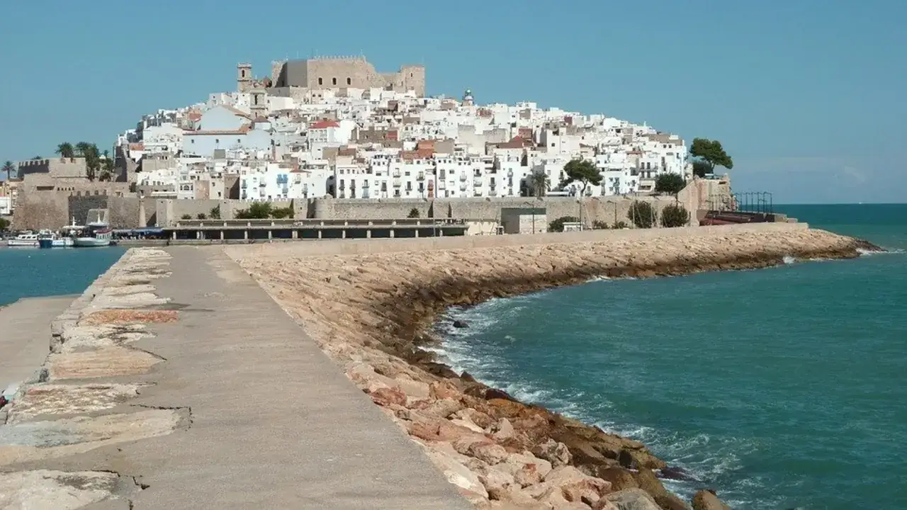 Pueblos costa Azahar: un castillo domina el caserío blanco apilado en una colina, con el mar turquesa rompiendo contra un rompeolas de rocas.