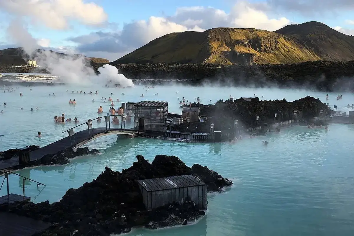 Ludzie kąpią się w turkusowej wodzie geotermalnego **blue lagoon** otoczonej czarnymi skałami i parującą mgłą.