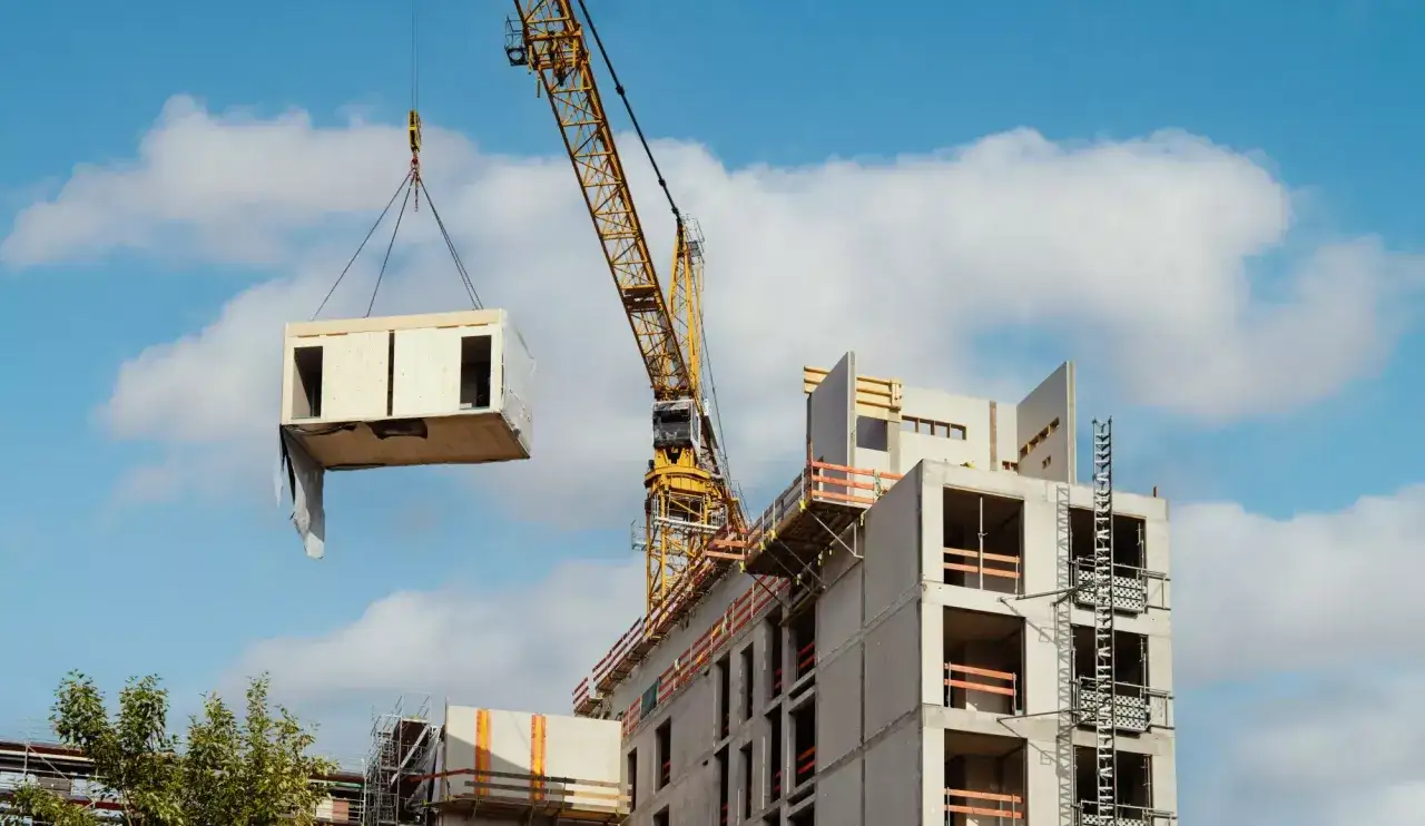 Une grue jaune soulève un module de maison modulaire en bois vers un bâtiment en construction.