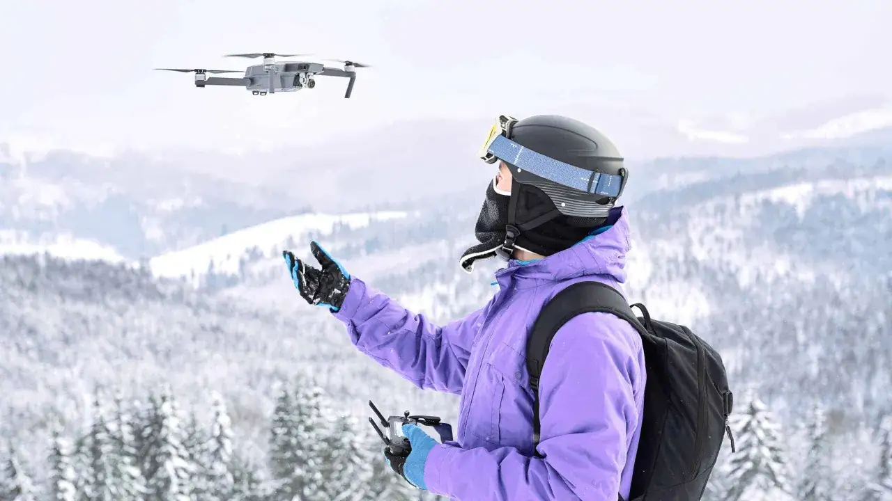 A person in ski gear controls a flying drone in cold, snowy weather, capturing the winter landscape.