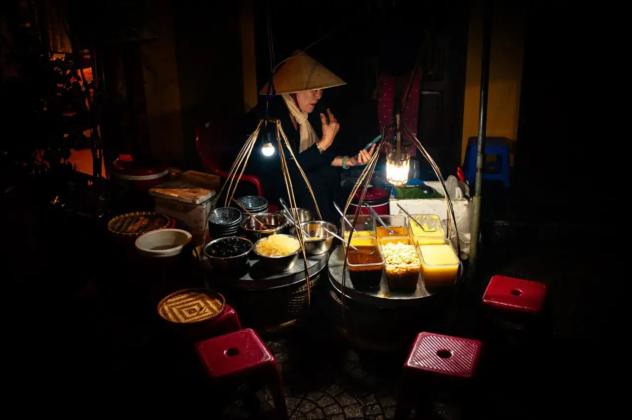 Photographie de voyage : une marchande de desserts asiatiques, sous un chapeau conique, consulte son téléphone à la lueur d'une lanterne.