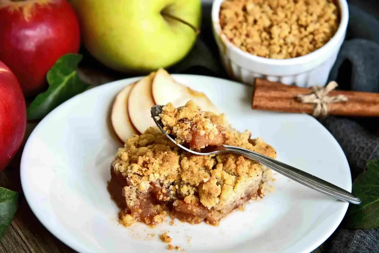 Une cuillère prélève un morceau d'un délicieux crumble pomme, avec des pommes fraîches et de la cannelle en arrière-plan.