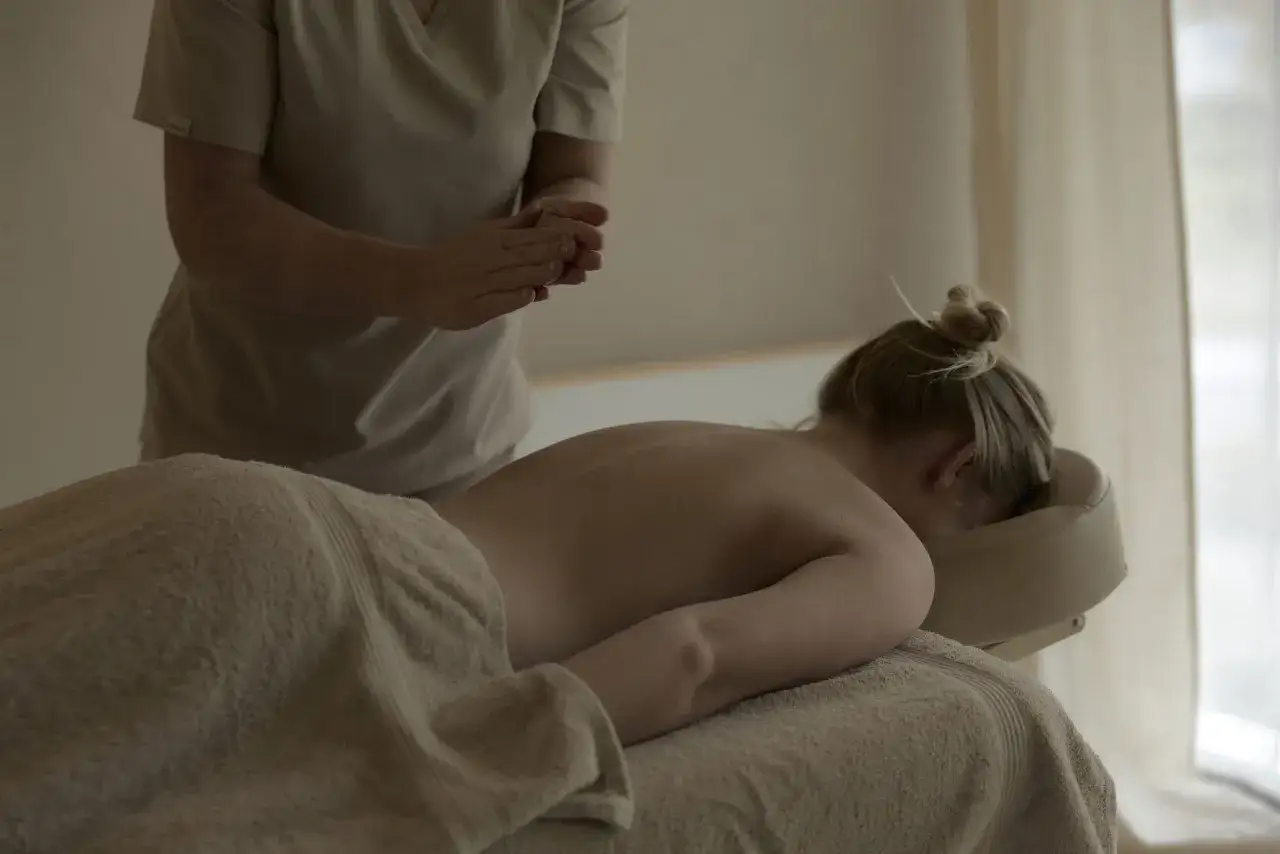 Terapeuta przygotowuje dłonie do masażu relaksacyjnego. Kobieta leży na stole do masażu, otulona ręcznikiem.