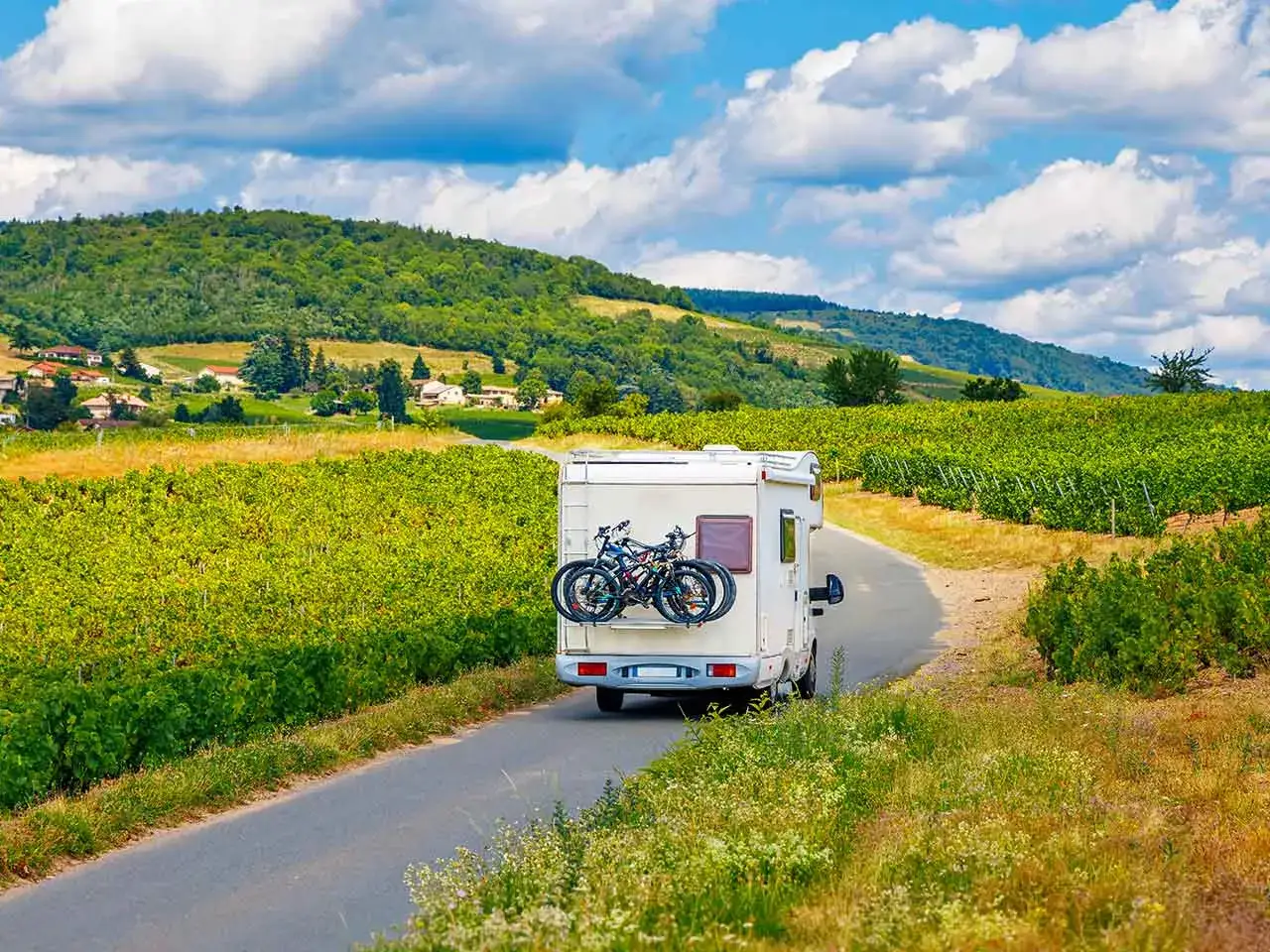 Un camper con biciclette sul retro percorre una strada tra rigogliosi vigneti, con colline verdi e un cielo azzurro punteggiato di nuvole. Un'avventura in **Toscana in camper**.