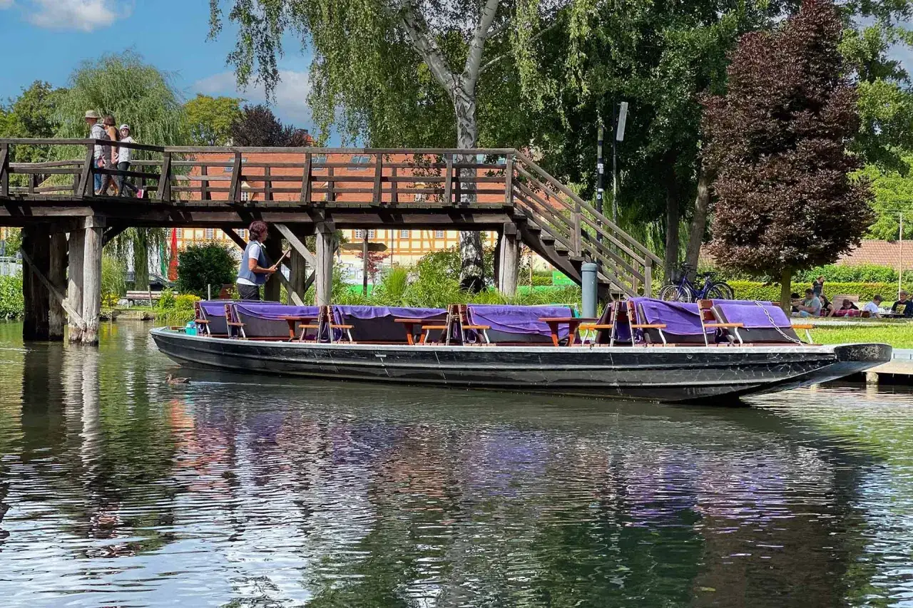 Kahnfahrt im Spreewald: Ein Ausflugsziel mit Charme. Ein Kahn mit violetten Bezügen wartet auf Gäste, während Menschen über eine Holzbrücke gehen.