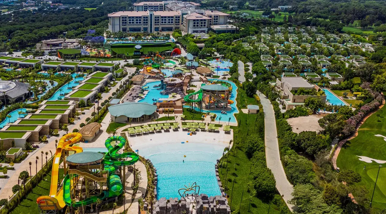 Ein weitläufiger Wasserpark mit Rutschen, Pools und Liegestühlen, umgeben von grünen Bäumen und luxuriösen Unterkünften. Das Regnum Carya bietet Spaß für die ganze Familie.