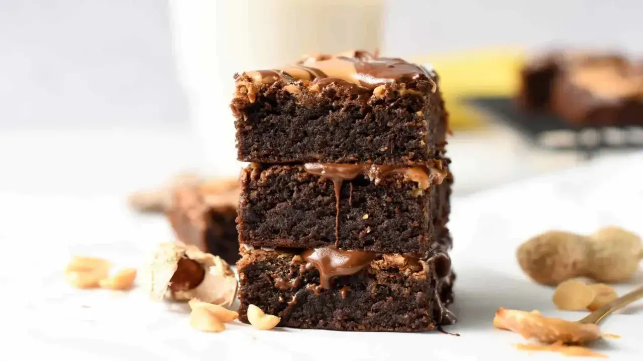 Stos trzech wilgotnych brownie z bananami i masłem orzechowym, polanych czekoladą. Obok leżą orzeszki ziemne.