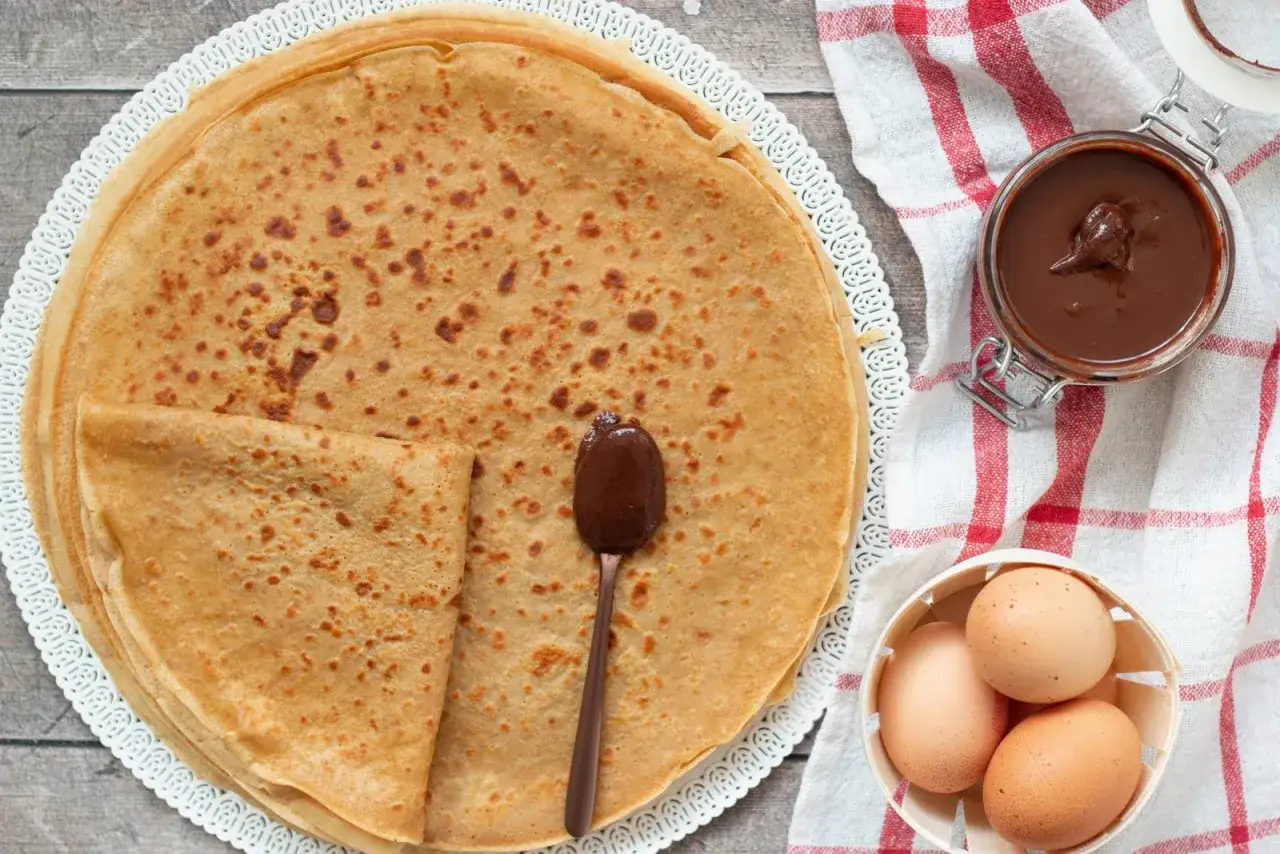 Pile de cr&ecirc;pes sans lactose, garnies d'une cuill&egrave;re de p&acirc;te &agrave; tartiner au chocolat. Des &oelig;ufs et un pot de p&acirc;te &agrave; tartiner sont &agrave; c&ocirc;t&eacute;.